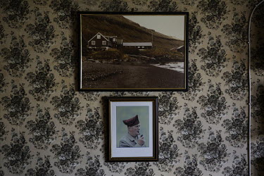 An old picture of a farm and a portrait of a Faroese man, wearing a traditional hat, hang in the living room of a farm.