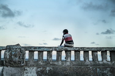 A boy sits alone on a wall beside the sea in the city's old town.